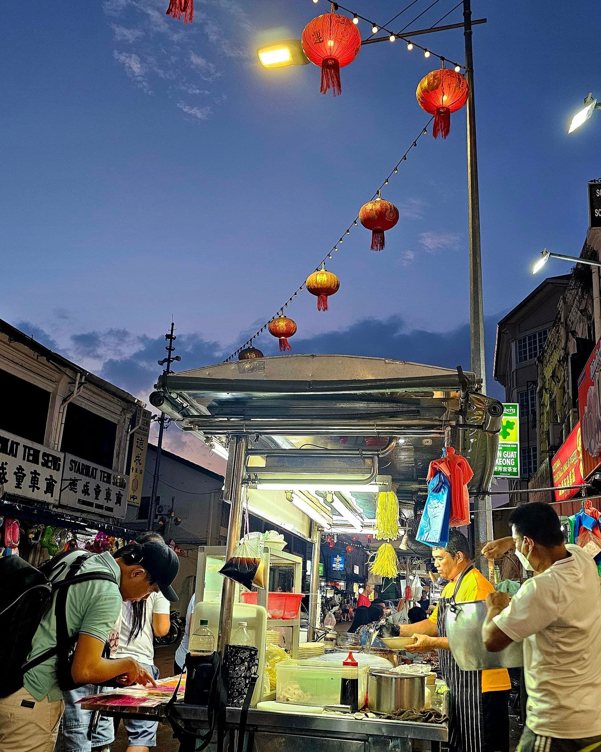 A vendor serving from a hawker cart under strung lanterns in George Town at dusk